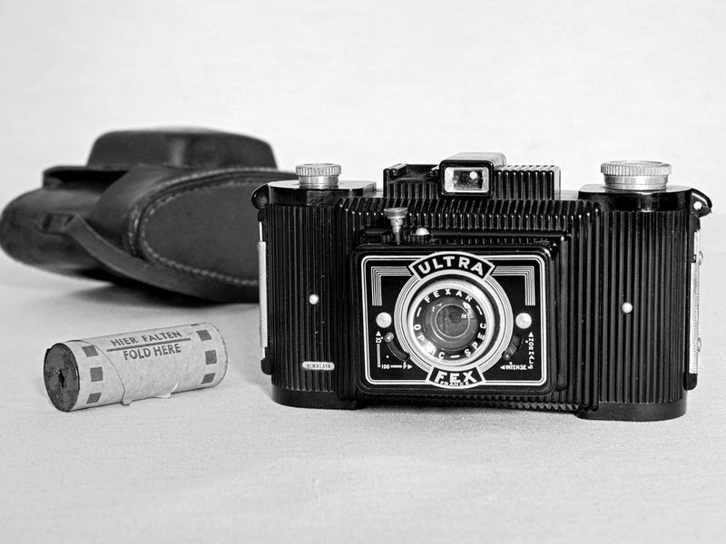 A black box camera beside its leather case and a roll of film