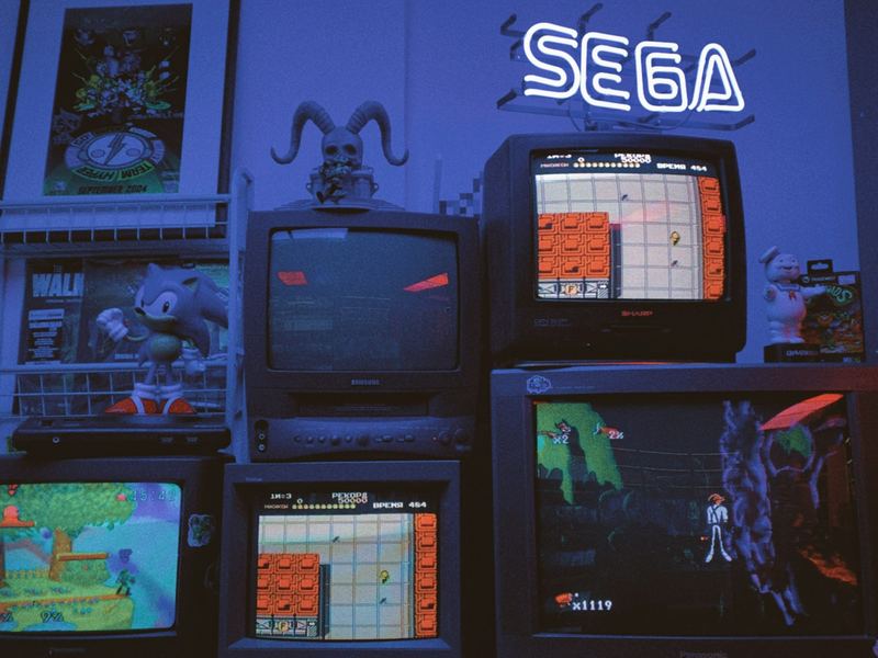 Stacked old TVs showing retro video games under a neon Sega sign